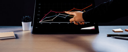 A professional workspace scene featuring a hand pointing at growth trends on a digital graph...