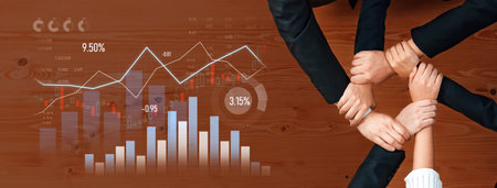 Hands of business professionals join together above a desk, surrounded by graphs and data,...