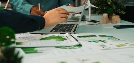 Team members engage in collaborative discussion, analyzing charts and graphs related to sustainability projects, showcasing a modern office with eco-friendly elements and greenery.の写真素材