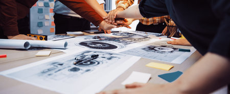 A dynamic creative team engages in a brainstorming session, hands together over design sketches and notes, showcasing collaboration and innovative ideas in an office setting.の写真素材