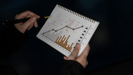 A close-up view of hands holding a notepad filled with graphs and charts, illustrating data trends for business analysis and financial planning in a professional setting.の写真素材