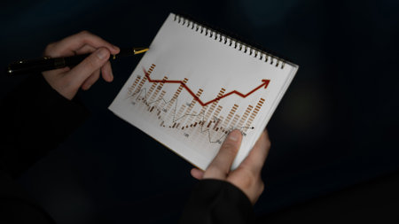 A focused business professional analyzes data trends in a dark setting, holding a notebook and pen, highlighting growth and financial insights.の写真素材