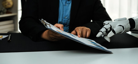 A human and a robotic hand collaborate over documents in a modern workspace, showcasing the integration of technology in business and innovation.の写真素材