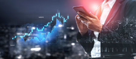 A business professional engages with a smartphone while analyzing charts and graphs of financial...