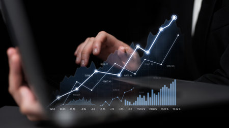 A businessman interacts with a laptop display, analyzing financial data and stock market trends through dynamic charts and graphs against a dark background.の写真素材