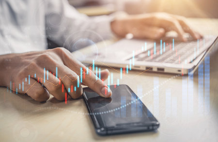 A business professional analyzes financial data using a smartphone and laptop. Rising stock charts...
