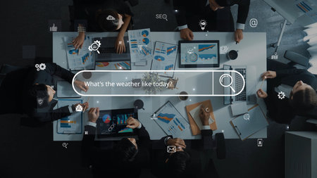 Aerial view of a diverse group engaged in a business meeting surrounded by laptops and charts. Digital icons emphasize collaboration in a modern workspace.の写真素材