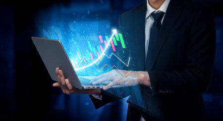 A business professional operates a laptop while analyzing dynamic financial graphs that represent...
