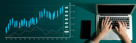 A top view of hands typing on a laptop with a smartphone nearby, showcasing financial graphs that...