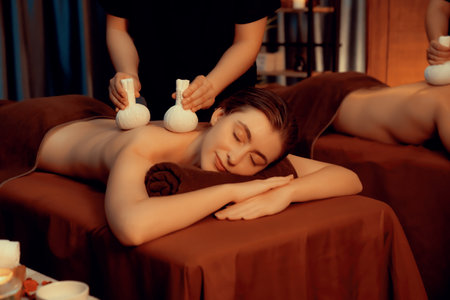 Hot herbal ball spa massage body treatment, masseur gently compresses herb bag on couple customer body. Serenity of aromatherapy recreation in warm lighting of candles at spa salon. Quiescentの写真素材