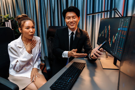 Asian businessman showing stock market application on mobile phone to partner pointing by pen,...