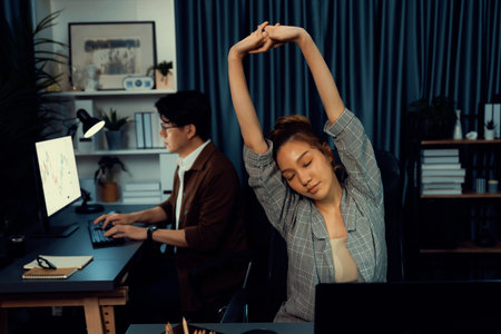 Businesswoman working on desk with stretching arm up and down manner with body health ache of strain overwork while coworker trading stock market on website on pc at late time.の写真素材