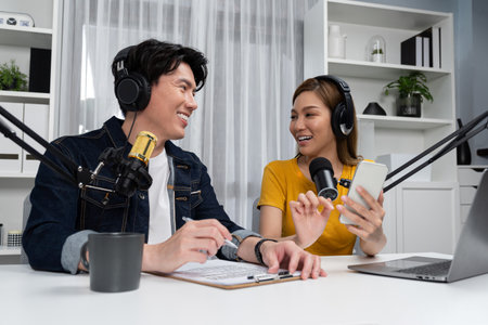 Smiling radio influencer hosts wearing headphones with channel social media online live streaming with talking script note with phone and laptop to listeners at morning at studio record. Infobahn.の写真素材