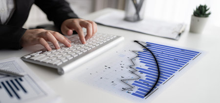 A business professional focuses on analyzing financial data at a modern office desk. The scene features a keyboard, charts, and a small plant, symbolizing productivity and innovation.の写真素材