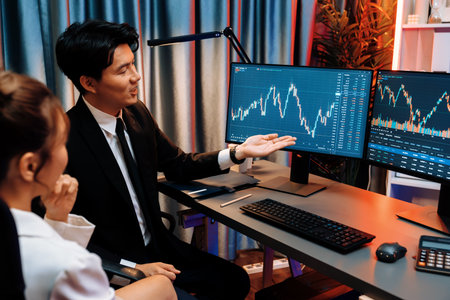 Asian businessman showing dynamic stock exchange currency rate statistic online to share with coworker, explaining to compare with high or low on computer screen at night neon modern office. Infobahn.の写真素材