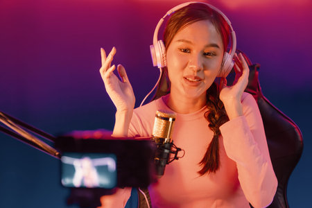 Host of creative broadcast listening to music on social media with listeners in a positive podcast, wearing pastel color headphones, using a microphone to record by camera at a neon studio.の写真素材