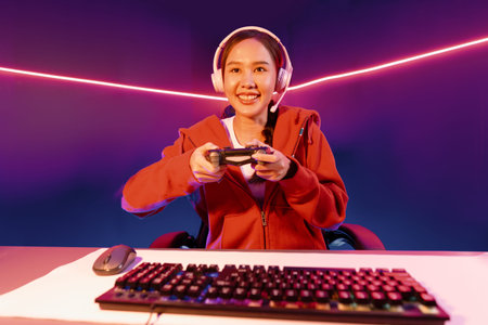 Host channel of smiling Asian girl streamer with joystick playing online game wearing headphones...