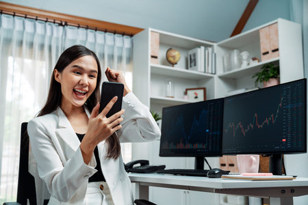 Successful young Asian businesswoman earning high profit raising fist up, looking smartphone for exchange stock markets financial technology with dynamic business graph at modern office. Stratagem.の写真素材
