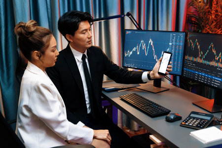 Asian businessman showing stock market application on mobile phone to partner pointing by pen,...