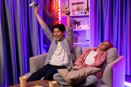 Sitting sofa of happy couple playing video game with joysticks, smiling man playing challenge, raising fist up to be winner the challenge at modern neon blub light living room at home.の写真素材