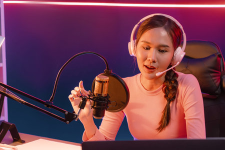 Smiling creative young beautiful Asian influencer talking in channel with positive podcast speech at...
