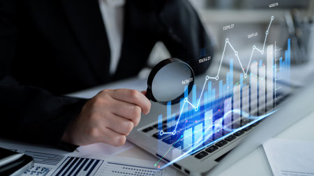 A businessman examines data trends using a magnifying glass on a laptop screen. The scene...