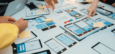 A vibrant workspace captures hands collaborating on a mobile app prototype. The scene features paper...