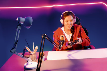 Host channel of beautiful Asian girl streamer with joystick playing online game wearing headphones...