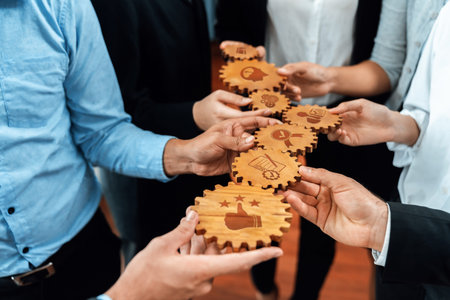 A group of diverse professionals joins hands, showcasing wooden gears with icons that represent teamwork and innovation, emphasizing collaboration and strategic planning.の写真素材