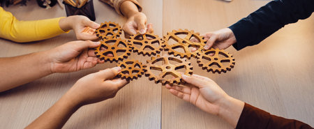 A group of diverse hands holds wooden gears in a collaborative workspace, symbolizing teamwork and...