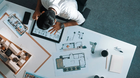 Top view of business man using tablet plan house design while sitting at meeting table with blueprint and equipment scatter around. Aerial view of civil engineer working at blueprint.の写真素材