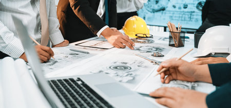 Professionals collaborate around a table full of engineering plans and modern technology,...