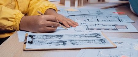 An architect focused on sketching detailed designs and plans at a work table filled with drawings,...