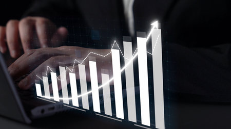 A business professional engages with a laptop while analyzing financial data trends represented...