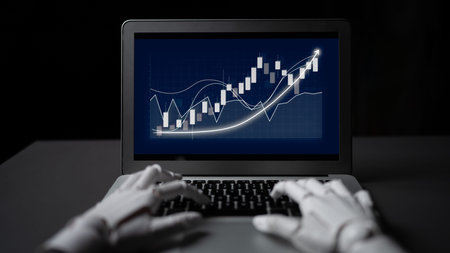 A robot hand interacts with a laptop screen showcasing financial market trends and analytics,...