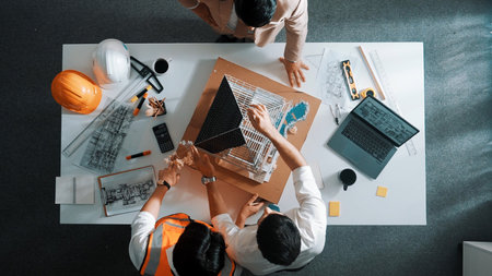 Top view of skilled architect designer team using pencil measuring house model. Group of civil engineer planning and designing blueprint while pointing at building at table with laptop.