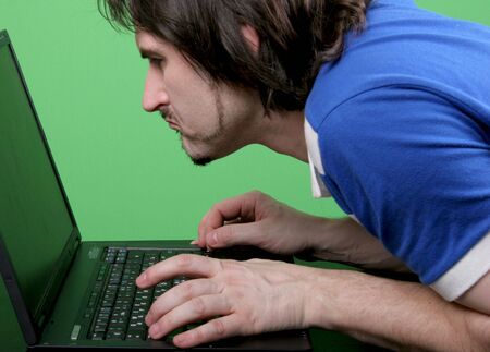 emotional man in blue t-shirt narrowly look to the display of deep gray laptop with green backgroundの写真素材