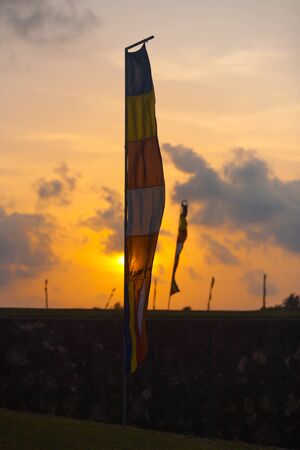 A colorful buddhist flag waves and obscures the setting sun on a bright orange sky in Galle, Sri Lankaの写真素材
