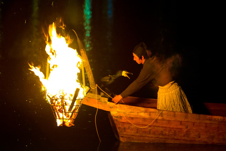 Inuyama, Japan - July 5, 2015: Japanese man in grass skirt, wooden boat, prepares a cormorant bird for ukai fishing drawing fish by a large flame at night on the Kiso River in Gifu Prefectureのeditorial素材