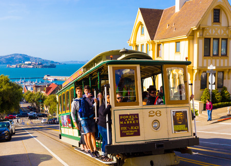 San Francisco, USA - May 12, 2016: Closeup of approaching Hyde Street cable car full of people standing on outside platform enjoying steep hill ride with Alcatraz Island backgroundのeditorial素材
