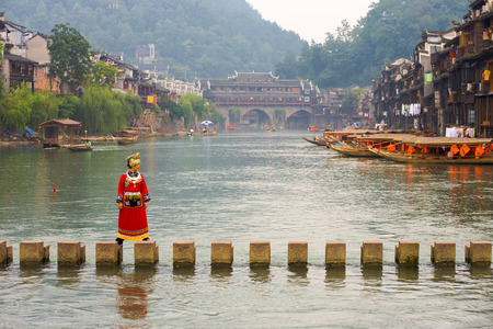 Fenghuang, China - September 12, 2007: A Chinese female tourist in traditional minority clothes crossing the stepping stone bridge across the Tuojiang Riverのeditorial素材
