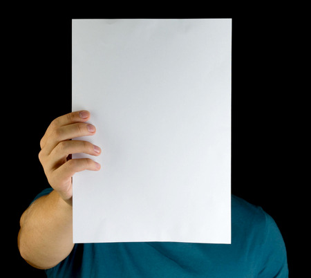 young man holding white blank paper vertically on extended arm copy space advertisement in front of his face isolated blackの写真素材