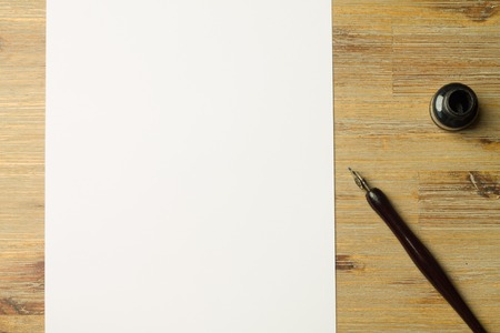 Fully visibe blank sheet of paper and pen on bright wooden office desk. Calligraphy pen and ink next to a fully visible sheet of paper with copyspace.の写真素材