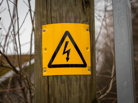 a yellow and black danger of death sign with triangle and lightening bolt on a wooden electric pole pylon outside resource with forest at the backgroundの写真素材