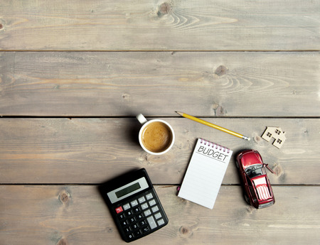 Notepad with budget handwritten on top of a wooden table with a miniature car and house symbolの写真素材