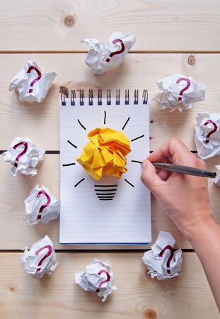 Drawing of a light bulb on a notepad with bright orange paper surrounded by crumpled pieces of paper with question marksの写真素材