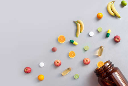 Vitamin pills and capsules alongside apples, bananas, lime and oranges coming out of a medicine jar, nutrition conceptの写真素材