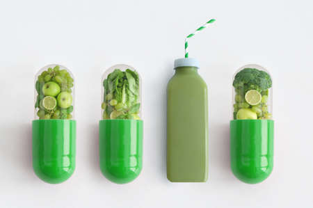 Green smoothie beverage alongside vitamin capsules containing fresh green fruits and vegetablesの写真素材
