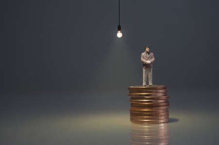 Miniature man standing on top of a pile of coins with light bulb shining aboveの写真素材