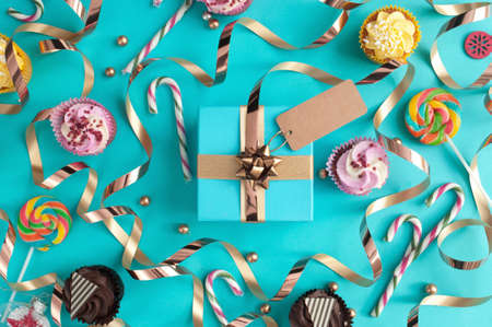 Gift box wrapped in gold ribbon with greeting card, surrounded by sweet treats including cupcakes and candyの写真素材